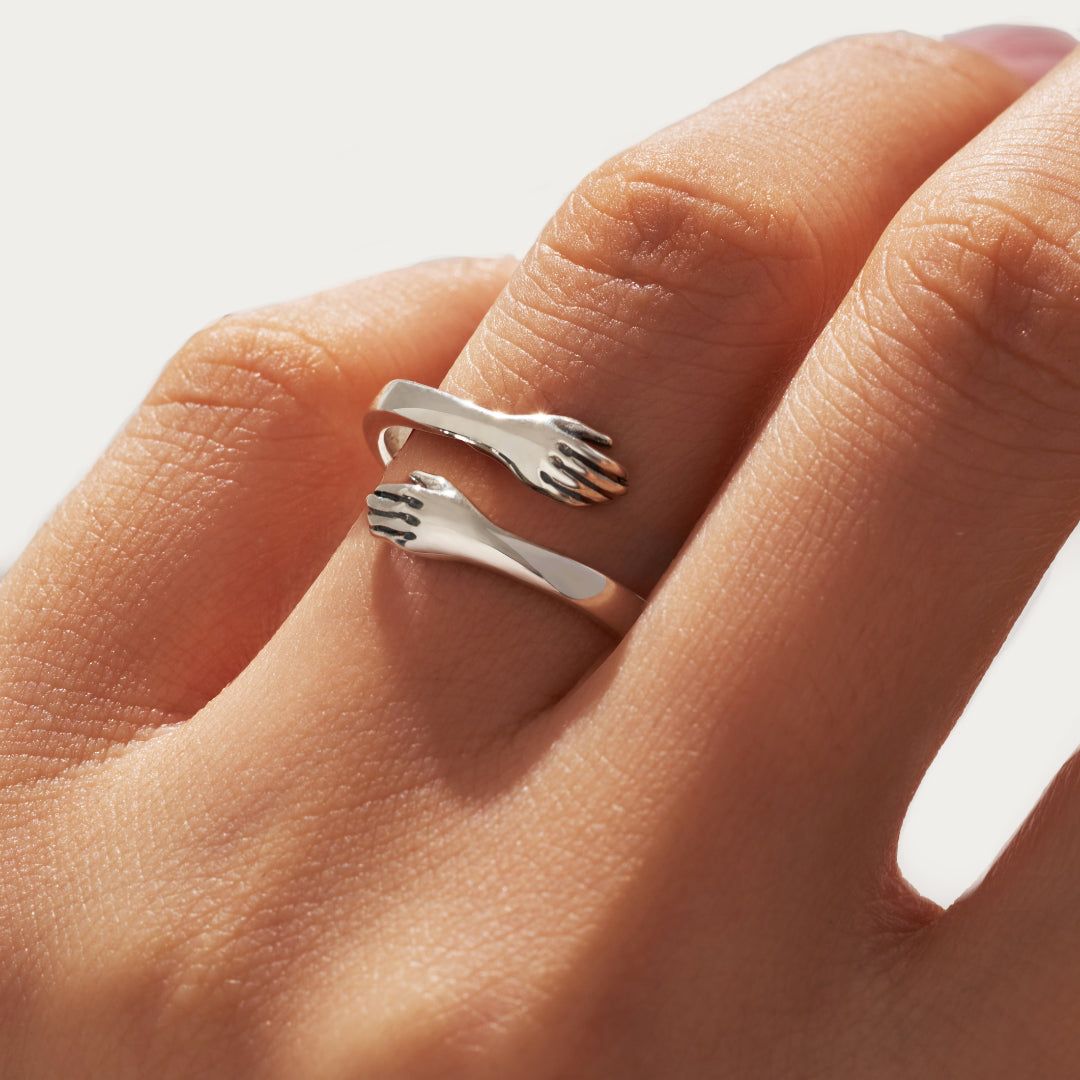 A person's finger adorned with the adjustable silver ring designed as two hugging hands.